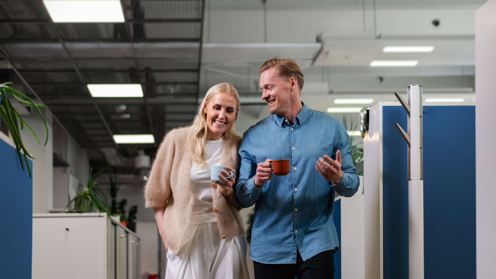 Two smiling colleagues, a woman and a man in casual business attire, walk and chat through a bright open-plan office holding takeaway coffee cups past desks and partitions.