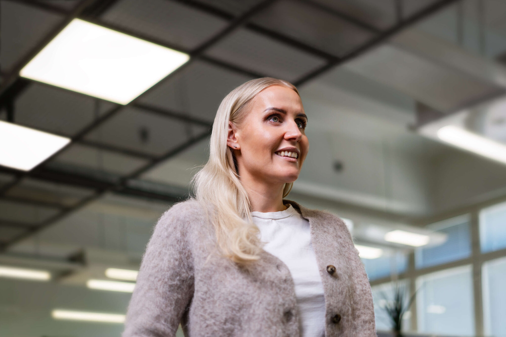 Hymyilevä vaalea nainen toimistotilassa katsoo ylös, päällään valkoinen paita ja vaalea neuletakki, taustalla kirkkaat suorakaiteen muotoiset kattovalot ja lasiseiniä.